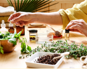 A table with fresh vegetables, green leaves, small bottles of oil, and a person in yellow clothing reaching out to the bottles, symbolizing natural digestive health support.